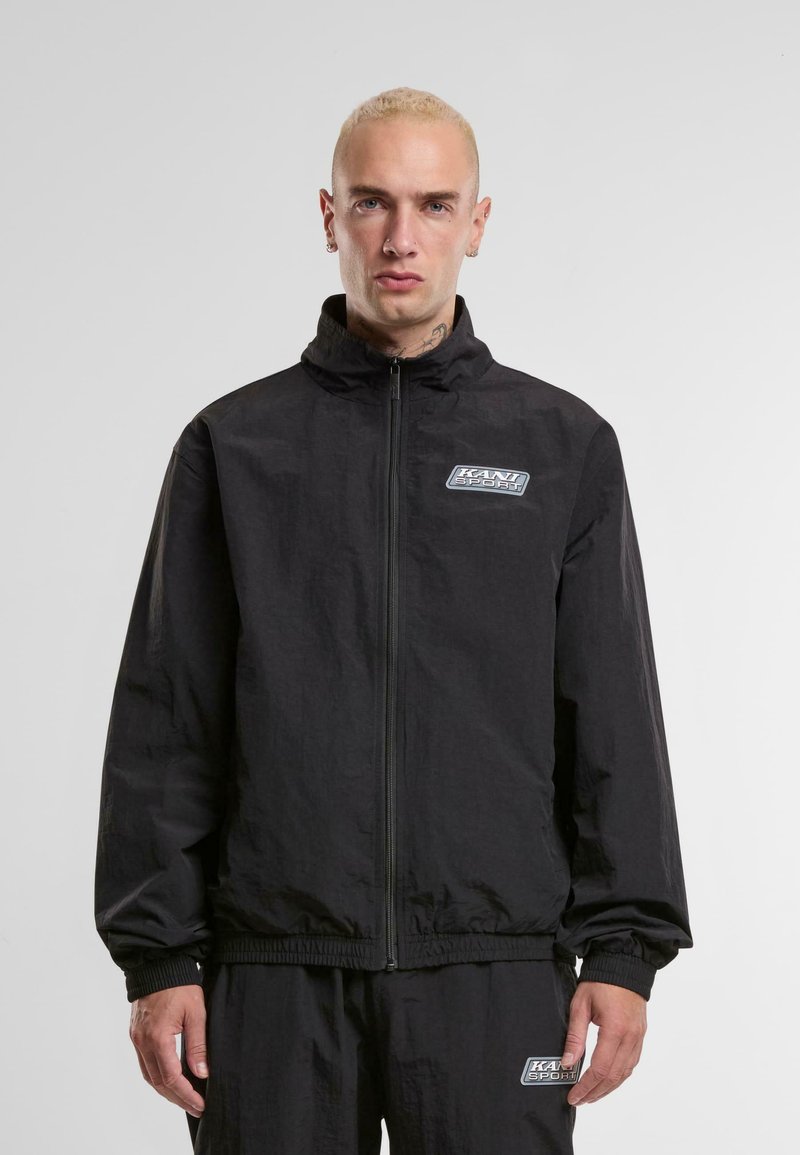 Black windbreaker jacket with a high collar, front zipper, and elastic cuffs. Features a logo patch on the chest and a textured fabric.