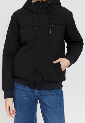 Person iført en sort lynlåstrøje med hætte og snore og blå jeans, med den ene hånd i lommen, mod en ensfarvet baggrund.