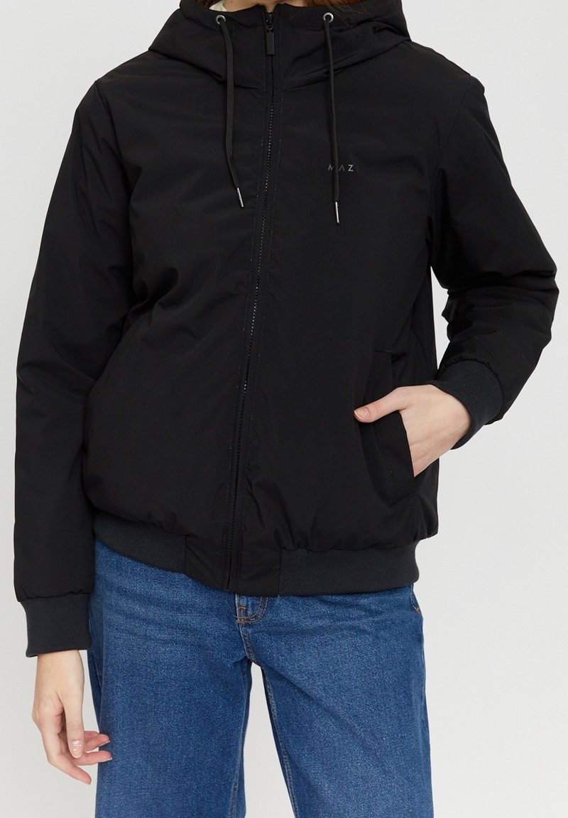 Personne portant une veste à capuche noire zippée avec des cordons et un jean bleu, une main dans la poche, sur un fond uni.