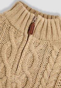 Beige kötött pulóver magas gallérral, kábelmintázattal, fél cipzárral és barna bőr díszítéssel. Texturált, bordás kialakítás.