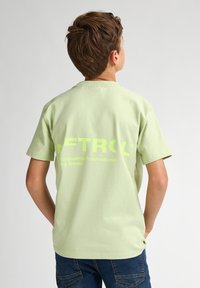 Světle zelené bavlněné tričko s krátkým rukávem. Na zadní straně je velký, tučný, neonově žlutý text a logo. Jednoduchý střih, kulatý výstřih, volný střih.