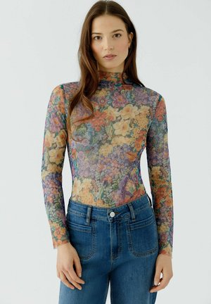 Frau trägt ein durchsichtiges, langärmeliges, geblümtes Oberteil, das in hoch taillierte blaue Jeans gesteckt ist, steht vor einem schlichten hellen Hintergrund.