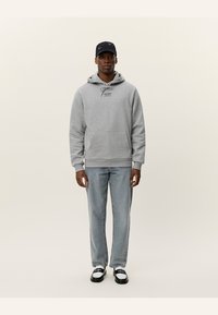 Grijze hoodie met voorzak en trekkoord, lichte spijkerbroek en zwarte pet. Model in zwart-witte schoenen staat tegen een lichte achtergrond.
