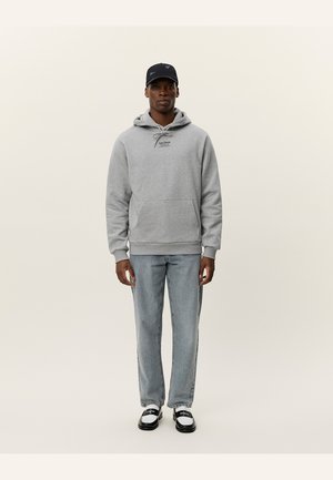 Grau Hoodie mit Fronttasche und Kordelzug, helle Jeans und schwarze Mütze. Model in schwarz-weißen Schuhen steht vor einem hellen Hintergrund.
