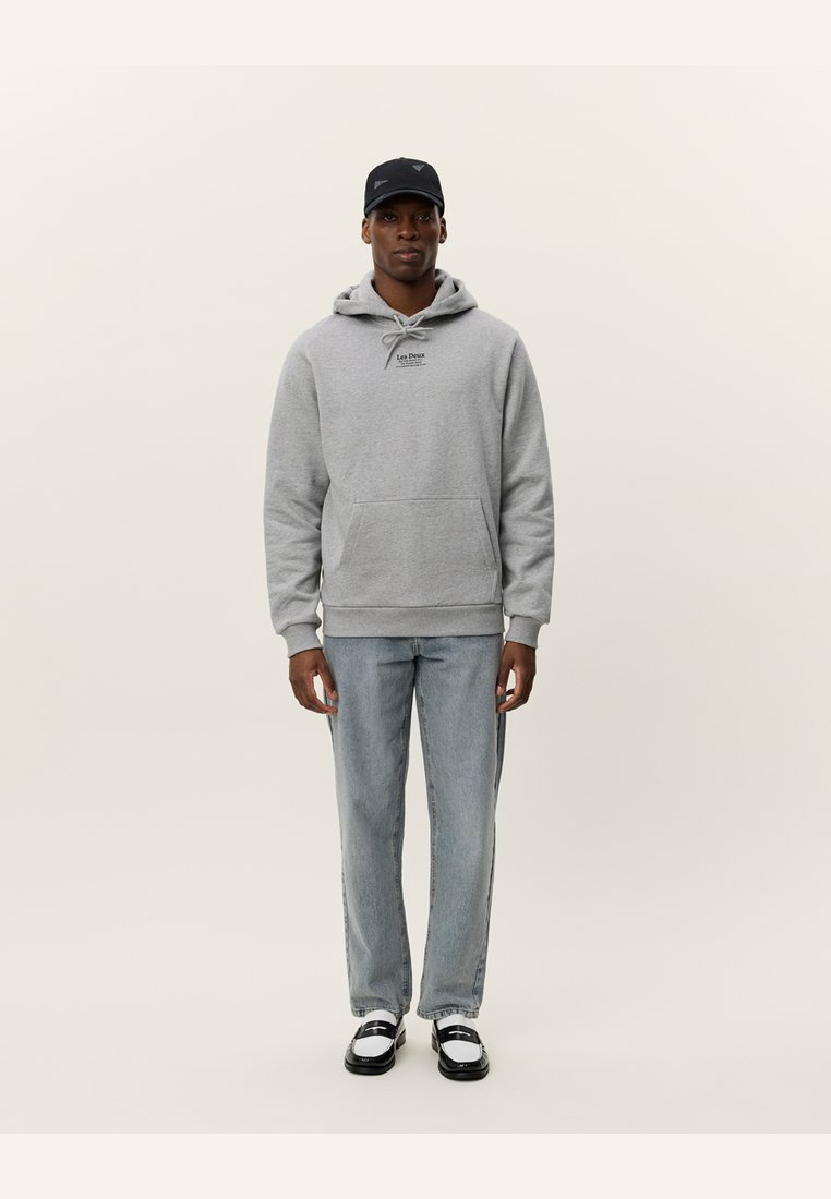 Grijze hoodie met voorzak en trekkoord, lichte spijkerbroek en zwarte pet. Model in zwart-witte schoenen staat tegen een lichte achtergrond.