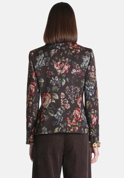 Femme aux cheveux bruns et lisses portant un blazer sombre à motif floral et un pantalon en velours côtelé marron, vue de dos sur un fond uni.