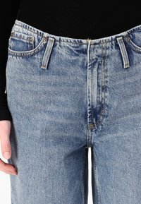 Lyseblå denimjeans med høy midje. Funksjoner inkluderer beltesløyfer, frontlommer og kontrasterende oransje søm.