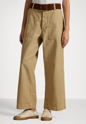 Beige bukser med vide ben og store forlommer, båret med et brunt bælte, hvid top og beige sneakers med gummisåler.