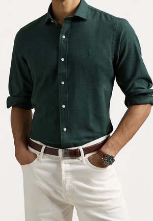 Homme portant une chemise boutonnée vert foncé avec manches retroussées, pantalon blanc, ceinture en cuir marron et montre-bracelet au cadran vert. Mains dans les poches.