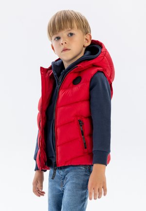 Rote gepolsterte Weste mit Kapuze, ausgestattet mit einem runden schwarzen Logo und zwei seitlichen Reißverschlusstaschen. Getragen über einem navyblauen Hoodie und Jeans.