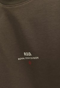 Hnědé bavlněné tričko s kulatým výstřihem. Na přední straně je nápis „R.D.D.“ a „ROYAL DENIM DIVISION“ v bílé barvě, s malou červenou tečkou.