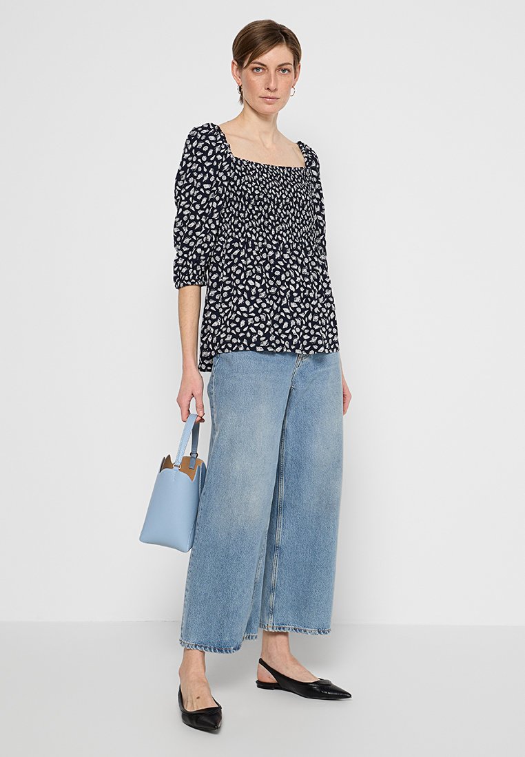 Marinebluse mit Blumenmuster und Puffärmeln, kombiniert mit hellblauen, weit geschnittenen Jeans und schwarzen, spitzen Ballerinas. Hält eine hellblaue Handtasche.