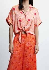 Korte mouwen, roze bloemenblouse met een geknoopt voorpand. Gecombineerd met feloranje wijde broek met bloemenpatroon voor een gecoördineerde look.