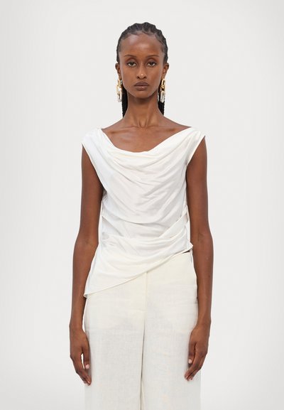 White draped blouse with asymmetric neckline and soft, textured fabric, paired with light-colored wide-leg pants.