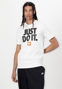 Balta medvilninė marškinėliai su dideliu juodu grafiniu tekstu "TIESIOG DARYK TAI." ir oranžine Nike logotipu. Apvalus kaklas, trumpomis rankovėmis, laisvas kirpimas.