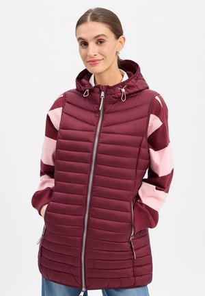 Frau mit braunen Haaren im Pferdeschwanz, die eine kastanienbraune gesteppte Kapuzenweste über einem rosa und kastanienbraun gestreiften Pullover trägt, mit den Händen in den Taschen.