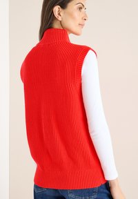 Rode geribbelde mouwloze trui met een hoge kraag, voorzien van een geribbelde zoom en daaronder een glad wit langermig shirt.