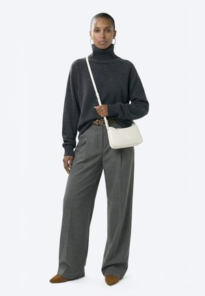 Femeie purtând pulover gri închis cu guler înalt, pantaloni largi gri, pantofi maro, curea cu imprimeu leopard și geantă albă de umăr, stând pe fundal alb.