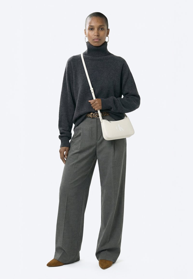 Žena u tamnosivom puloveru s visokim ovratnikom, širokim sivim hlačama, smeđim cipelama, pojasom sa leopardovim uzorkom i bijelom torbom preko ramena, stoji ispred bijele pozadine.