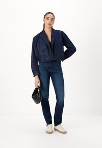Veste en denim bleu marine coupée avec des boutons double boutonnage, deux poches avant, assortie à un jean foncé et des baskets beiges, tenant un sac noir.