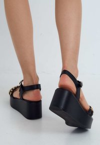 Sandali platform neri con tacco spesso, cinturino alla caviglia regolabile e dettaglio a catena decorativa sulla parte anteriore. Texture liscia e design minimalista.