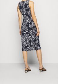 Kvinna som bär en marinblå ärmlös klänning i knälängd med vit blommig tryck och svarta klacksandaler med genomskinliga blockklackar.