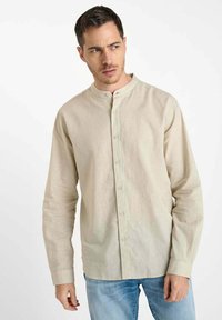 Man in een beige mandarin kraag button-up shirt en blauwe spijkerbroek, die naar links kijkt tegen een effen witte achtergrond.