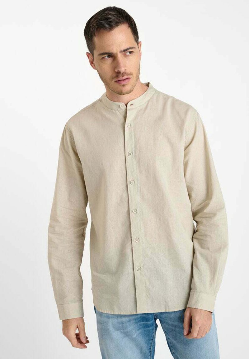 Man in een beige mandarin kraag button-up shirt en blauwe spijkerbroek, die naar links kijkt tegen een effen witte achtergrond.