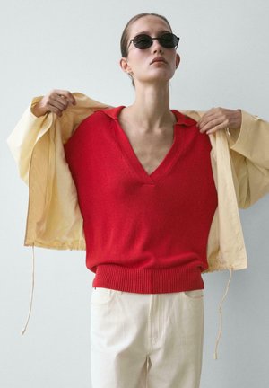 Person wearing black sunglasses and a red v-neck knit sweater removing a light beige jacket, standing against a plain light background.