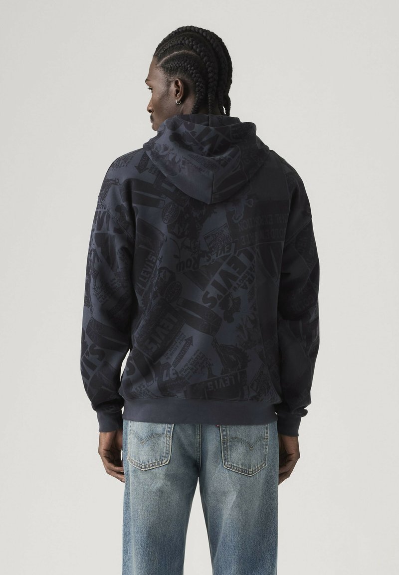 Man with braided hair wearing a dark printed hoodie and light blue jeans, standing facing away against a plain light background.