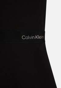 Melns Calvin Klein jostasvērts ar gludu tekstūru, ar baltiem logotipa burtiem modernā fontā uz eleganta auduma virsmas.