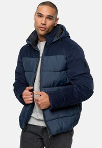 Puffy navy jacket with a hood, featuring corduroy accents and smooth panels, zip front, and ribbed cuffs, worn over a gray sweatshirt.
