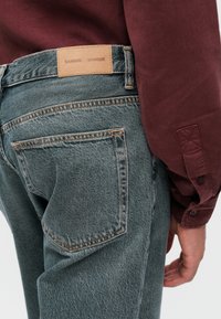 Gai pelēki denima džinsi ar oranžiem diegiem, ādas zīmola lapa jostas vietā un redzamām aizmugurējām kabatām. Stilizēti ar tumši purpursarkanu kreklu.