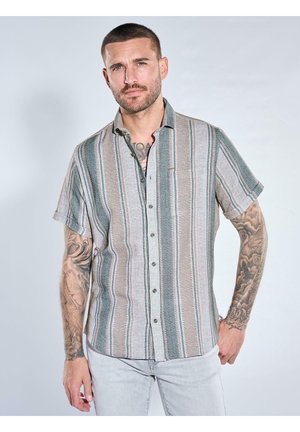 Mann mit kurzen Haaren und Tattoos trägt ein kurzärmliges, gestreiftes Hemd mit Knöpfen und hellgraue Hose vor neutralem Hintergrund.