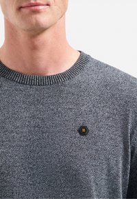 Grijze gebreide trui met een geribde ronde hals en een zwart hexagonaal logo-opdruk aan de linkerkant. Zachte textuur, casual ontwerp.