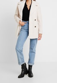 Kremowy, puszysty płaszcz o podwójnym zapięciu, z dwoma ciemnymi guzikami, noszony na czarnym, prążkowanym topie i jasnoniebieskich dżinsach z podwiniętymi mankietami, z czarnymi botkami sięgającymi kostek.