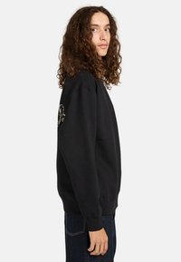Felpa nera con collo rotondo e maniche lunghe, presenta un design testurizzato sulla manica sinistra, indossata con jeans blu scuro.