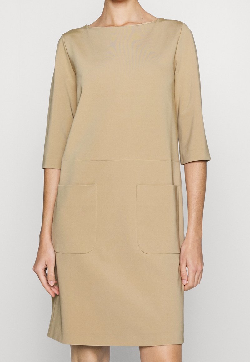 Robe beige longueur genoux avec manches trois-quarts et deux grandes poches avant, portée par une personne les bras détendus le long du corps.