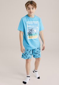 Ljusblå bomulls t-shirt med grafiken "VACAY TIME" och palmträddesign, i kombination med matchande badshorts med svarta palmträdsmönster.