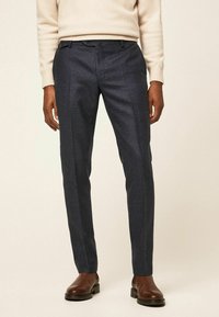 Tmavě modré tailored kalhoty z texturované látky, s plochým předním dílem, dvěma bočními kapsami a jemnými švy, doplněné hnědými botami.
