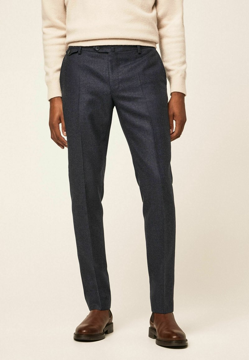 Tmavě modré tailored kalhoty z texturované látky, s plochým předním dílem, dvěma bočními kapsami a jemnými švy, doplněné hnědými botami.