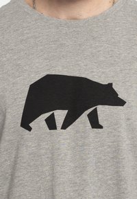 Camiseta gris con un gran gráfico de la silueta de un oso negro centrado en el pecho. La tela parece suave con una textura sutil.