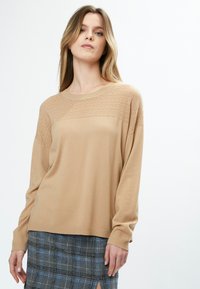 Beige gestrickter Pullover mit strukturierter Passe, langen Ärmeln und gerippten Bündchen. Kombiniert mit einem grauen karierten Rock mit blauen Akzenten.