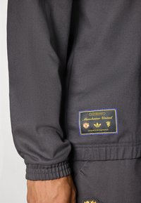 Pilkas sportinis kostiumas su ribotu rankogaliu, su pleistru, kuriame matomi „Manchester United“ ir „Adidas“ logotipai, auksiniai ir mėlyni akcentai bei tekstūruota medžiaga.