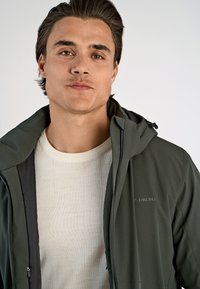 Tummanvihreä takki hupulla, vetoketjukiinnityksellä, hienovaraisella brändäyksellä, ja vaalea beige neulepaita sen alla, esillä valkoista taustaa vasten.