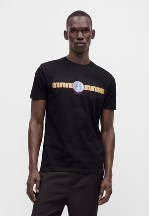 Mann trägt schwarzes T-Shirt mit gelbem und rotem Versace-Logo und Medusa-Kopf-Design, steht vor schlichtem hellem Hintergrund.