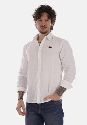 Uomo con capelli ricci che indossa una camicia bianca a maniche lunghe con bottoni e jeans scuri, in piedi con una mano in tasca e tatuaggi visibili sulle braccia.