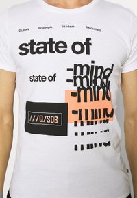 Bílý bavlněný tričko s výrazným černým a broskvovým textem. Grafika obsahuje fráze "stavu" a "mysli", s různými písemnými styly a rozvrženími.