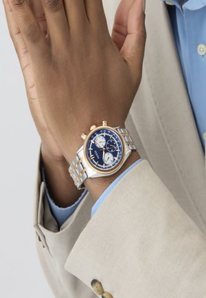 Montre en acier argenté et doré avec un cadran bleu, trois sous-cadrans et des fonctionnalités de chronographe. Conçue avec un bracelet en maillons métalliques.