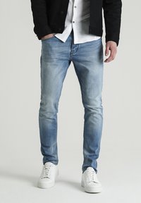Jeans da uomo azzurri in una vestibilità slim con aree sbiadite. Abbinati a una camicia bianca e un giubbotto nero, e sneaker bianche. Design semplice.